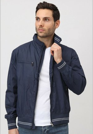 Veste zippée bleu marine avec un col montant, en tissu texturé, poignets rayés et poche poitrine, associée à un t-shirt blanc et un jean.