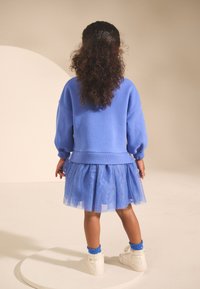 Kék pulóver púpos ujjakkal, réteges kék tüllszoknyával párosítva. Fehér tornacipő és kék fodros zokni egészíti ki a szettet.