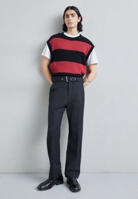 Gilet en tricot noir et rouge à rayures porté sur un t-shirt blanc, associé à un pantalon noir taille haute et des mocassins noirs. Design minimaliste.