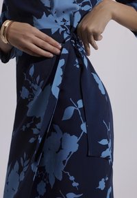 Granatowa sukienka w kwiatowy wzór, z węzłem w talii i zwiewnym materiałem. Gładka tekstura i długie rękawy widoczne. Złota bransoleta jako dodatek.