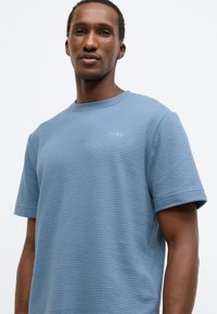 Kék rövid ujjú póló texturált szövettel és kerek nyakkivágással. A mellrészen kis "HUGO" logó hímzéssel díszített.