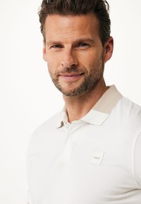 Uomo con capelli castani corti e barba corta che indossa una polo bianca con colletto beige e un piccolo logo quadrato "MEXX" sul petto.