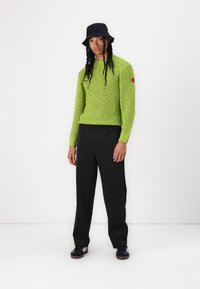 Sweater verde de malha com uma trama solta e aberta; chapéu bucket preto; calças pretas ajustadas; ténis pretos com riscas brancas. Acento vermelho na manga.