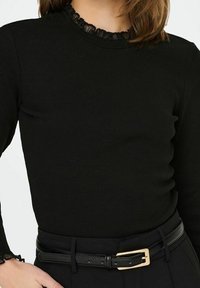 Svart ribbad långärmad topp med en volangkrage och handledsmuddar, kombinerad med ett svart bälte med en guldfärgad spänne.