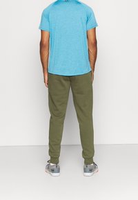 Man i blå kortärmad träningsskjorta, olivgröna joggingsbyxor och grå sneakers som står mot en enfärgad vit bakgrund.