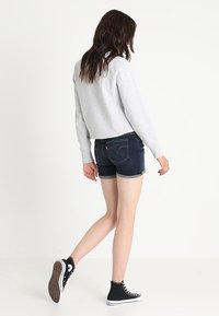 Grijze cropped sweatshirt, donkere denim short met omgeslagen zoom en zwarte hoge sneakers, gezien van achteren.