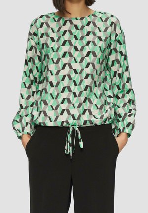 Femme portant un haut à manches longues à motifs avec des formes géométriques vertes, noires et grises et un pantalon noir avec les mains dans les poches.