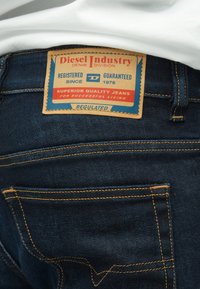 Temno modre kavbojke iz jeansa z vidno etiketo iz rjavega usnja. Etiketa ima rdeče in modre poudarke ter rumene šive na žepih.