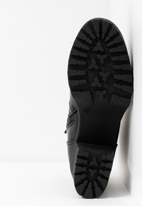 Semelle de botte noire avec un motif profond faisant face à la caméra, botte positionnée debout contre un mur blanc d'angle.