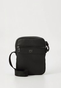 Sort lædertaske med tekstureret yderside, lynlås, justerbar rem og præget logo foran. Kompakt rektangulær form.