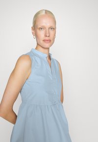 Vestido sem mangas azul claro com uma pala de botões e colarinho mandarim. Apresenta cintura franzida e uma textura de tecido lisa e suave.