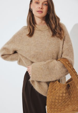 Donna che indossa un maglione peloso beige e pantaloni scuri, che tiene una grande borsa in pelle intrecciata color cuoio su uno sfondo semplice.