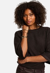 Pull noir en tricot à manches larges, porté avec plusieurs bracelets en or. Le mannequin a les cheveux bouclés et se tient les bras croisés.