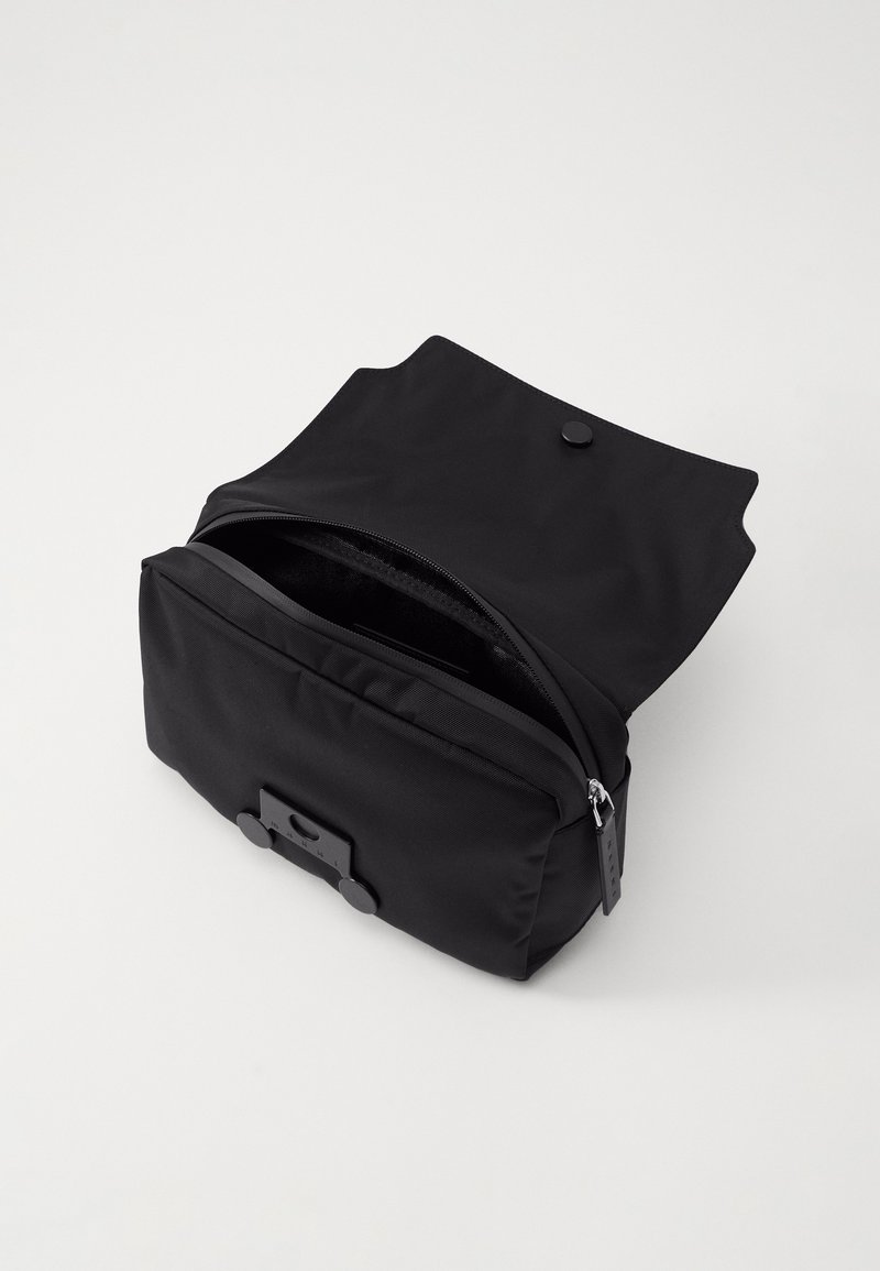 Borsa nera in nylon con chiusura a pattina, dotata di scomparto con cerniera e bottone a pressione. Design compatto con una superficie liscia e ferroni minimali.