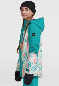 Teal og mønstrede skijake med hætte, prydet med pink og blå abstrakte designs. Sort hue og sorte handsker supplerer outfittet.