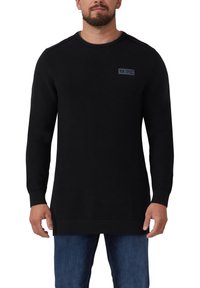 Homme portant un long pull en tricot noir texturé avec un petit écusson sur la poitrine et un jean bleu, se tenant devant un fond blanc.