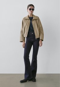 Ljusbrun kort jacka med avslappnad passform, kombinerad med mörka utsvängda jeans och svarta solglasögon. Slät textur med minimal detaljering.