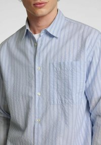 Camicia azzurra chiara con sottili righe verticali di blu scuro, colletto corto e taschino a sinistra. Tessuto morbido con una vestibilità casual.