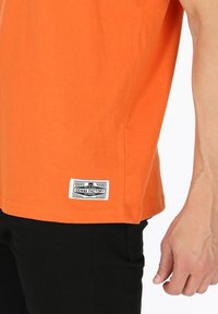 Oranssi puuvillainen t-paita lyhyillä hihoilla, jossa mustavalkoinen kudottu etiketti helman kohdalla. Suora pinta, rento istuvuus.