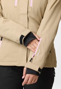 Beige jakkeærme med pink lynlås og justerbar velcrolukning, hånd delvist inde i ærmelommen, sort inderlag synligt ved håndleddet.