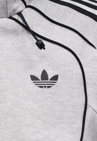 Ανοιχτό γκρι φούτερ με μαύρο λογότυπο Adidas trefoil, μαύρα κορδόνια με πλαστικό στοπ και μαύρες ρίγες κατά μήκος της ραφής του ώμου.