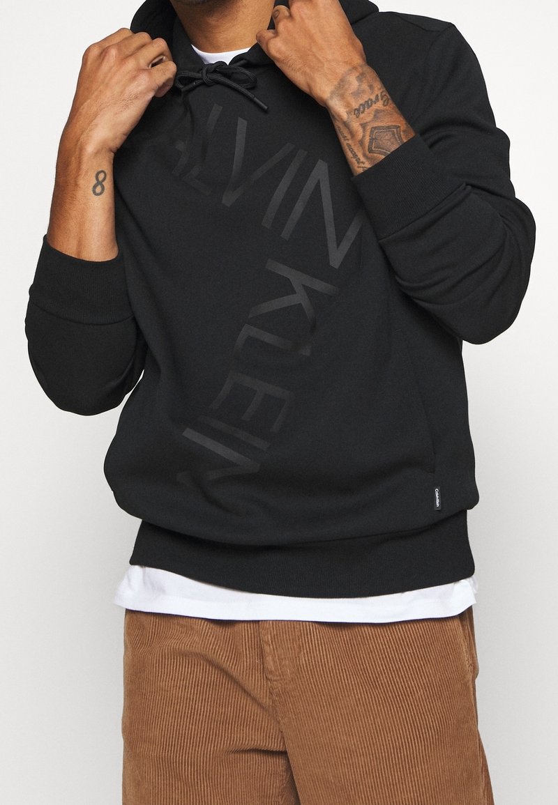Homme portant un sweat à capuche noir Calvin Klein avec cordons, un sous-maillot blanc et un pantalon en velours côtelé marron, montrant des tatouages sur les deux avant-bras.