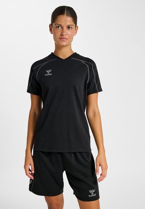 Kvinde iført sort Hummel sportstøj, stående med afslappede arme, kiggende let til siden mod en ensartet lys baggrund.