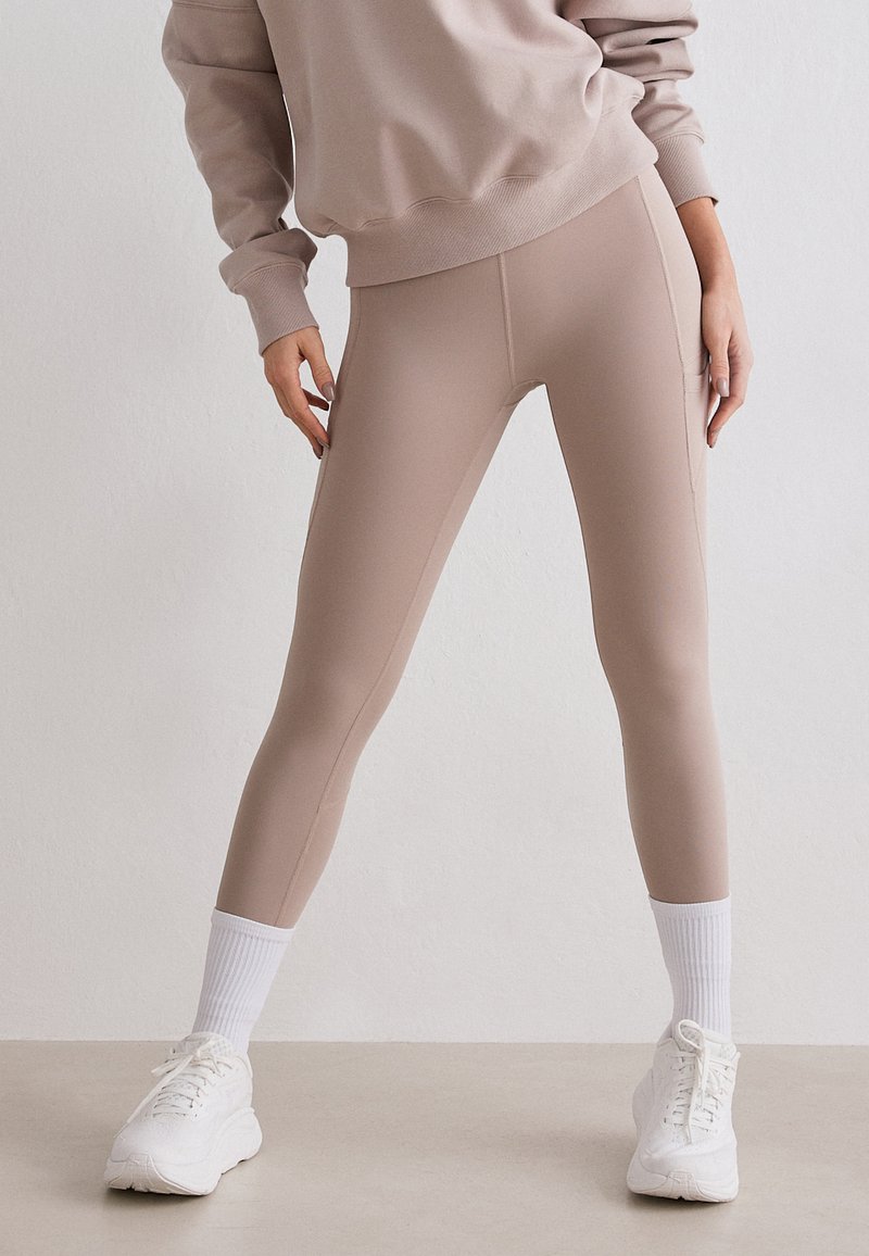 Bas du corps d'une personne portant des leggings beiges, un sweat assorti, des chaussettes blanches et des baskets blanches épaisses, debout contre un mur clair uni.