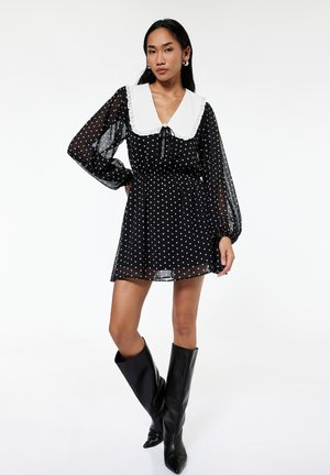 Donna che indossa un vestito nero trasparente a pois con un grande colletto bianco e stivali neri al ginocchio, in piedi con una mano sui fianchi.