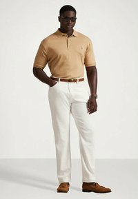 Άνδρας που φοράει μπεζ πόλο μπλούζα, λευκά παντελόνια, καφέ ζώνη και μπαλαρίνες, με γυαλιά ηλίου και ρολόι, που στέκεται σε έναν απλό φόντο.