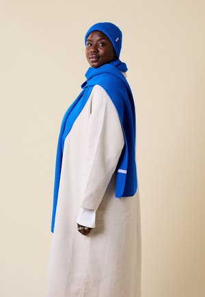 Blauer Strickmütze und Schal kombiniert mit einem langen beigen Kleid. Der Schal legt sich über die Schultern und weist eine gerippte Textur und weiße Akzente auf.