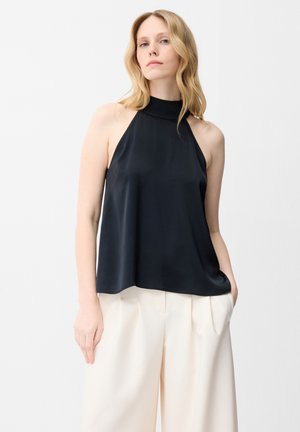 Femme aux cheveux blonds porte un haut noir sans manches col halter et un pantalon blanc ample, debout devant un fond blanc uni.