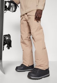 Pantalons de snowboard beige à coupe décontractée, avec des poches zippées et un logo sur la cuisse, associés à des bottes de neige noires.