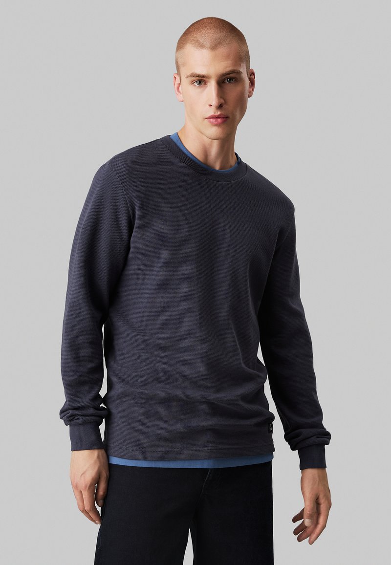 Felpa a maniche lunghe color blu navy scuro con collo rotondo e superficie testurizzata, indossata sopra una camicia azzurra chiara, abbinata a pantaloni scuri.