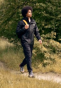 Svart vattentät jacka och byxor med elastiska manschetter, kombinerat med grå och orange vandringsskor. Individu bär en ryggsäck med gul rem.
