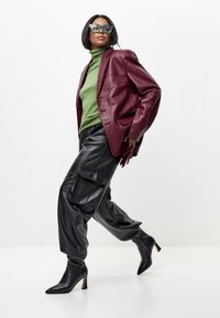 Mujer con un blazer de cuero burdeos oversized, suéter de cuello alto verde, pantalones cargo de cuero negros, botas de tacón negras y grandes gafas de sol verdes caminando.