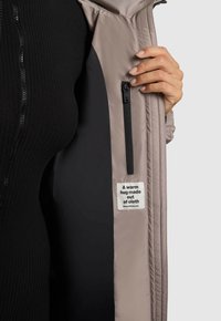 Svetlo béžová vypchávková bunda s vreckom na zips, vystavená s čiernou podšívkou a štítkom so slovami "Teplé objatie z látky."