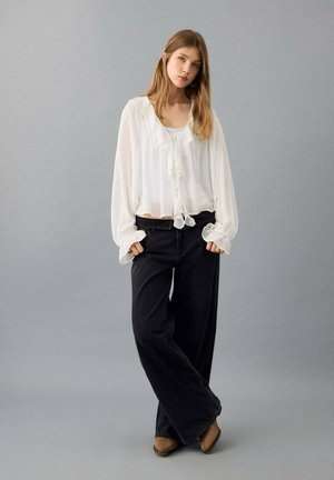 White blouse with ruffled details, tied at the waist, paired with black wide-leg jeans. Model stands confidently against a gray background.