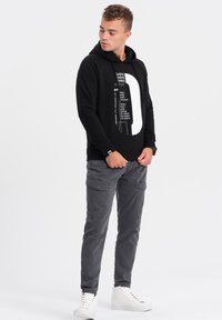 Sort hættebluse med hvid grafisk tekst, parret med grå cargo-bukser. Modellen har hvide højtop sneakers på. Glat stof og afslappet pasform.