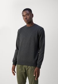 Sweat-shirt gris foncé avec un col rond et des manches longues, présentant un tissu texturé et un logo subtil sur la poitrine gauche. Associé à un pantalon vert.