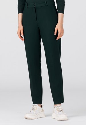 Femme portant un pantalon noir sur mesure et des baskets blanches épaisses, debout avec les bras détendus le long du corps sur un fond uni.