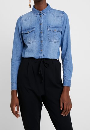 Persoon draagt een lichtblauw denimoverhemd met klepzakken en zwarte hooggetailleerde broeken met een trekkoord in de taille.