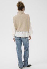 Beige knitted turtleneck sweater vest over a white blouse, paired with light blue loose-fitting jeans and black shoes.