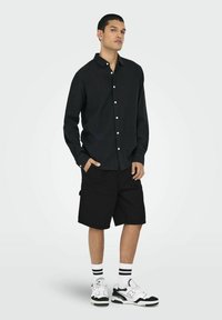 Zwarte overhemd met kraag, lange mouwen; gecombineerd met zwarte cargoshorts en witte sneakers met zwarte accenten en gestreepte sokken.