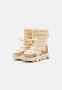 Beige polstrede støvler med høj skaft, elastiske stropper og en tekstureret sål. Har en buet bund og metalliske hardware detaljer.