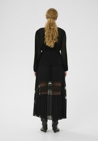 Čierne plisované maxi šaty s priehľadnými panelmi a dlhými rukávmi. Dizajn zahŕňa priliehavý živôtik a prúdiacu sukňu, doplnené o čižmy po kolená.