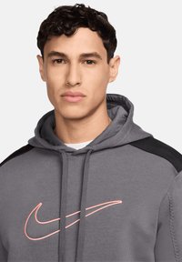 Felpa grigia con cappuccio e lacci, con dettagli neri sulle spalle e il logo rosa dello swoosh Nike sul petto. Tessuto morbido.