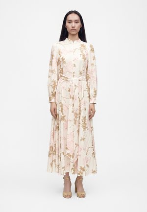 Femme portant une robe florale à manches longues, longueur cheville, avec un col montant et une ceinture nouée à la taille, associée à des talons ouverts beige.