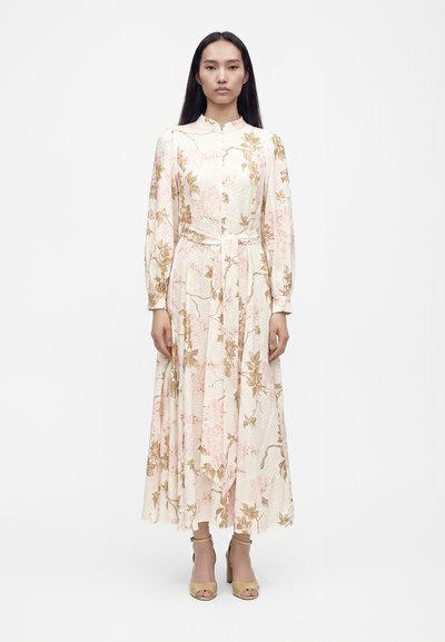 Kvinde iført en langærmet, ankellang blomsterkjole med højt krave og bindebælte i taljen, kombineret med beige åbne hæle.