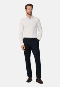 Lange mouwen polo in crème met een lichtblauwe kraag, gecombineerd met donkere broek. Model staat, handen in de zakken, met bruin veterschoenen aan.
